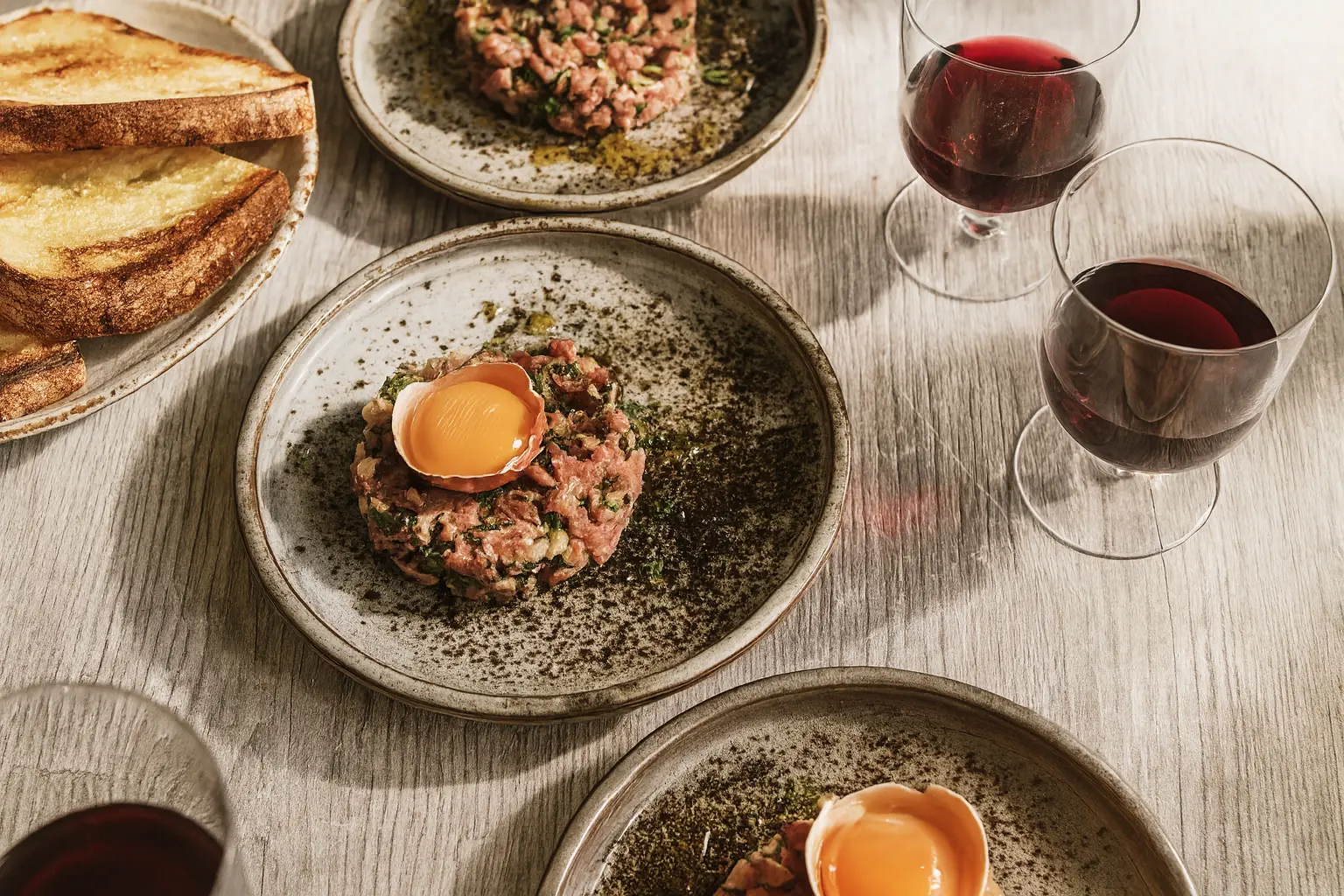 Artistically plated steak tartare topped with an egg yolk in its shell, alongside glasses of red wine and toast.
